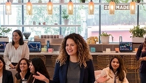 Un grupo de mujeres conversa y sonríe en un espacio de trabajo compartido con pantallas de gráficas al fondo.